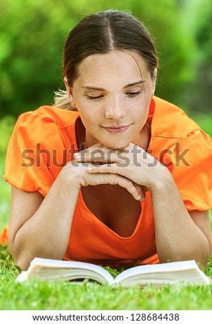 Dark-haired smiling beautiful young woman in orange blouse lying on grass and reading book, against green of summer park.