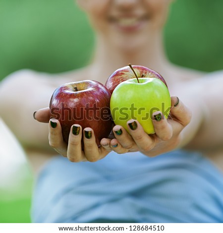 Beautiful young woman with bare shoulders stretches apple, against green of summer park.