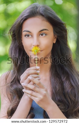 Beautiful smiling dark-haired young woman smelling yellow flower, against summer green park.