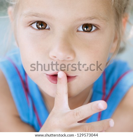 Beautiful little blonde girl with curly hair puts her finger to mouth, against background of summer city park.