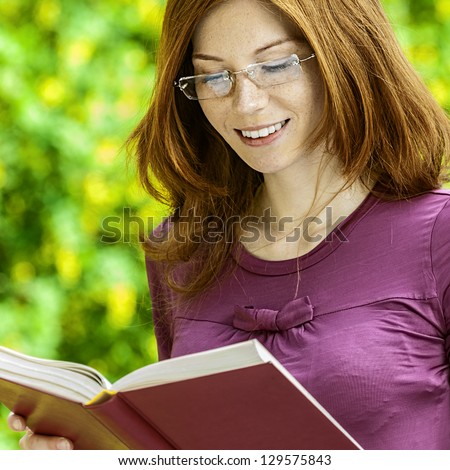 Red-haired smiling beautiful young woman with glasses reading book, against green of summer park.