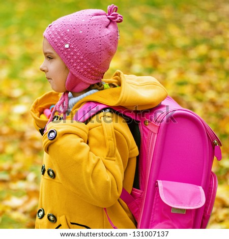 Little beautiful girl in yellow coat and pink backpack goes to school in autumn park.