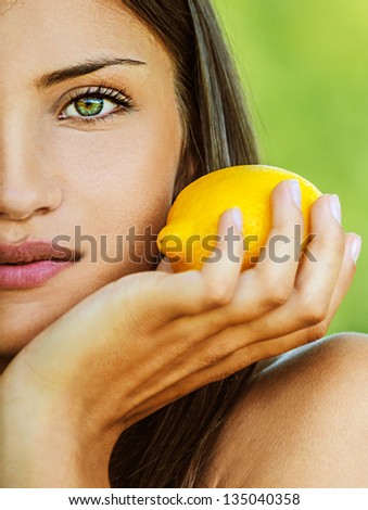 Portrait half of face young beautiful woman with bare shoulders holding lemon yellow, on green background summer nature.