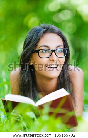Beautiful smiling dark-haired young woman lying on grass and reading red book, against summer green park.