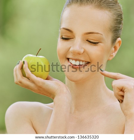 Beautiful young woman with bare shoulders points at apple and smiles, against green of summer park.