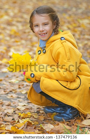 Beautiful smiling little girl in yellow coat collects fallen leaves in autumn park.