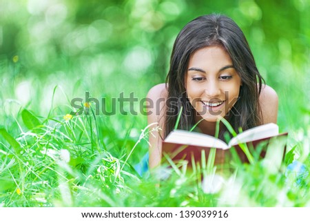 Beautiful smiling dark-haired young woman lying on grass and reading red book, against summer green park.