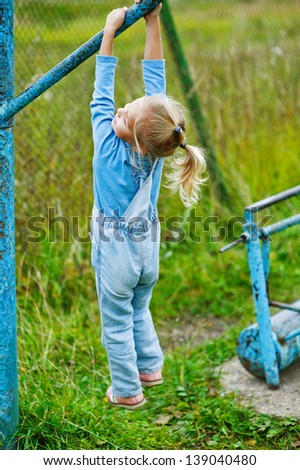 Beautiful little girl hanging on old exercise equipment in deserted park.
