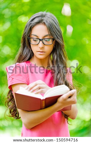 Young beautiful dark-haired woman in red dress and glasses holds book, against green summer garden.