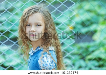 Portrait of beautiful smiling little girl with curly hair near fence of grid, against background of summer city park.