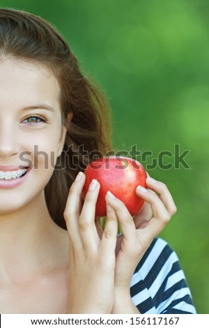 Portrait of young cheerful attractive smiling woman eating red ripe apple at summer green park.