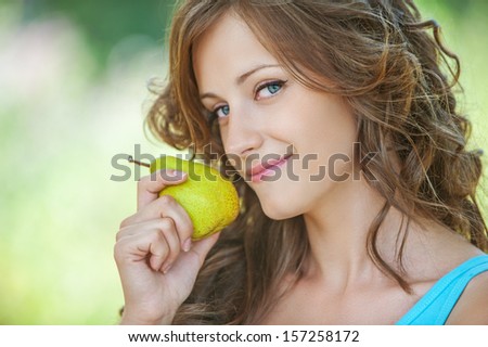 Close-up portrait of pretty smiling dark-haired freckled woman wearing blue t-shirt, holding juicy pear at summer green park.