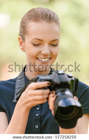 Beautiful smiling young woman in dark blouse photographs on camera, against green of summer park.