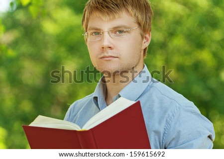 young, charming, serious student (male) with glasses reading book on background of summer park