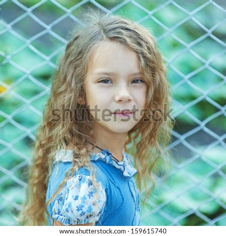Portrait of beautiful smiling little girl with curly hair near fence of grid, against background of summer city park.