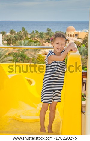 Beautiful little girl stands near large orange water park slides.