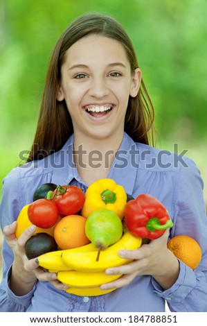 Smiling teenage girl holding banana, peppers, pears and oranges, against green of summer park.