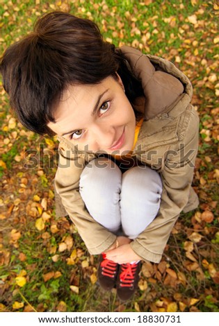 Beautiful dark-haired woman sits on earth covered with autumn fallen leaves.