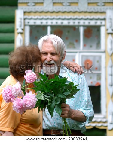 Elderly husband and the wife hold flowers in hands and laugh and joke against the house.