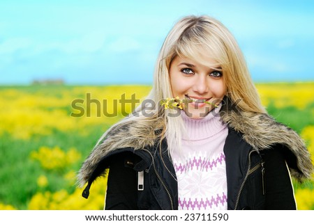 Young beautiful woman holds in  teeth a yellow floret against meadow and the sky