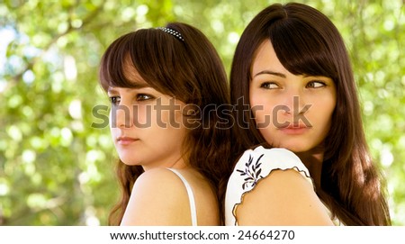 Two beautiful young women stand a back to a back in wood and look afar.