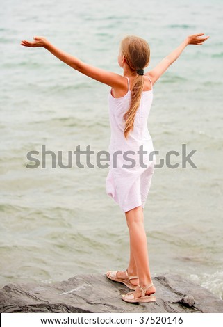 Young schoolgirl stands on seacoast and lifts hands towards to summer sun.