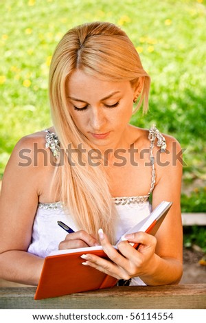 Beautiful smiling girl-student writes to writing-books against green summer nature.
