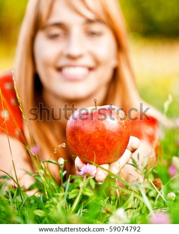 Beautiful smiling girl lies on lawn and gives red apple.