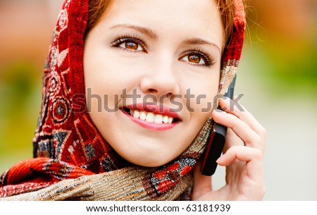 Young beautiful smiling woman in motley red headscarf talks on cellular telephone, against city structures.