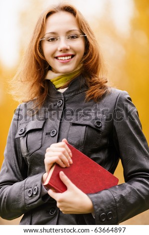 Young smiling girl-student in glasses has control over red book against yellow autumn nature.