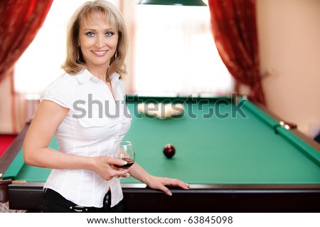 Beautiful mature smiling woman with wine glass in hands stands about billiard table in poolroom.