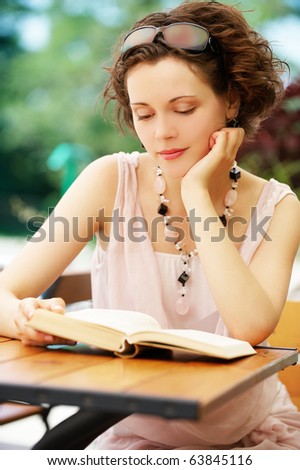 outside portrait of beautiful curly young woman sitting at the park cafe table and reading book