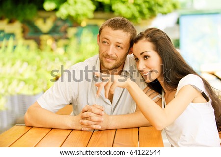 Young couple sit at little table in summer cafe. Girl shows to young man forefinger on something in distance.