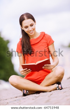 Young beautiful smiling woman in red dress sits in lotus pose on stone blocks and reads book.