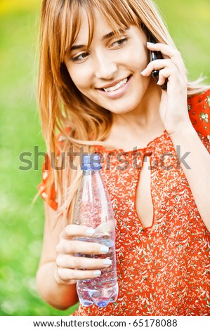Young beautiful student in red clothes speaks on cellular telephone and has control over bottle with water, on green summer background.