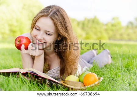Charming girl with apples lies on green grass and reads book.