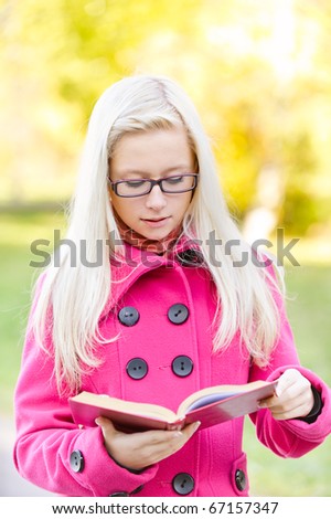 Beautiful fair-haired girl in pink coat reads red book, against autumn city park.