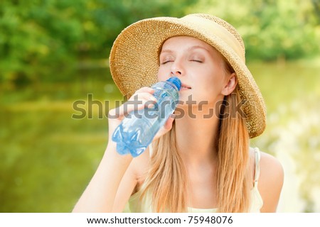 Young beautiful girl in straw hat against lake in city park drinks water from plastic bottle.