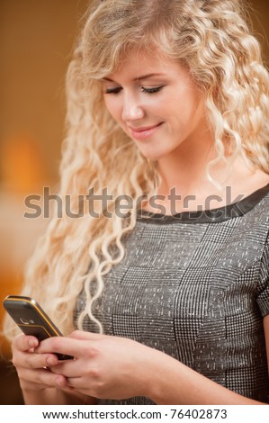 Beautiful curly-haired smiling girl sits at table and types text on mobile phone, against magnificent interior.