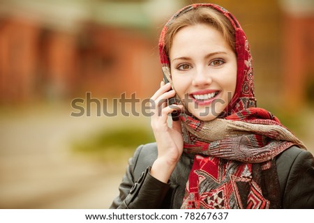 Young beautiful smiling woman in motley red headscarf talks on cellular telephone, against city structures.