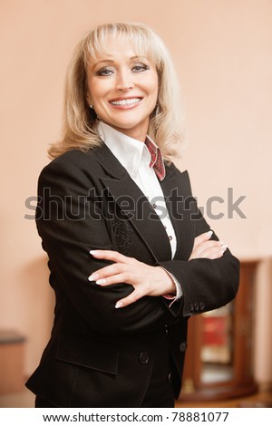 Portrait of beautiful smiling mature business woman in white shirt and jacket with crossed hands, against magnificent interior.