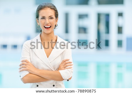 Young beautiful laughing business woman in glasses and with crossed hands against magnificent office.