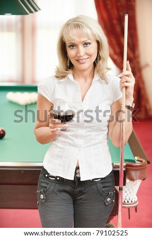 Beautiful mature smiling woman with wine glass in hands stands about billiard table in poolroom.