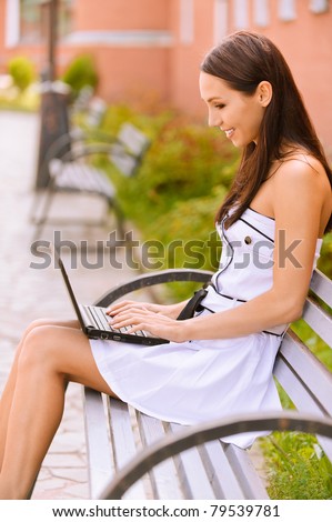 Young beautiful brunette student in profile sits on bench about university and works behind black laptop.