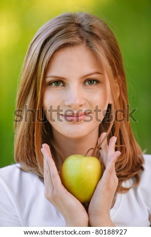 Young fine woman holds apple and smiles, on green background of summer city park.
