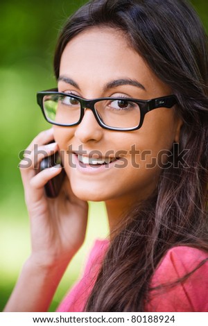 Young beautiful dark-haired woman in glasses black are right talks on cellular telephone and smiles, against green summer garden.