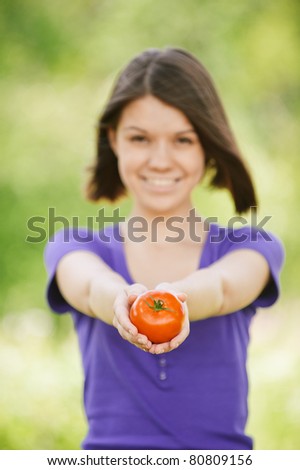 Portrait of young smiling cheerful brunette woman in violet blouse holding tomato at summer green park.