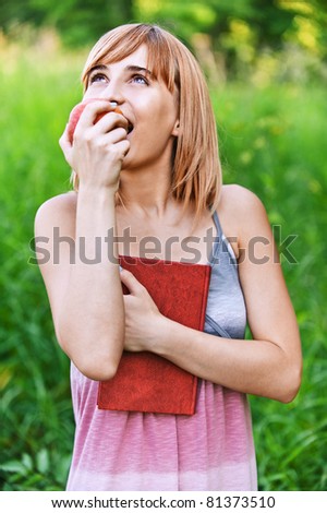 Portrait of young attractive woman with red book eating apple and looking to sky at summer green park.