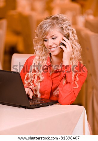 Young charming smiling woman-student works on black laptop and speak on mobile phone against beautiful interior.