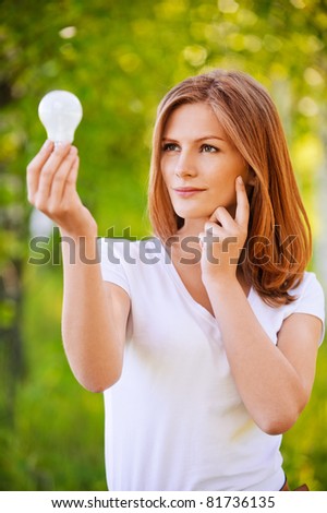Portrait of young beautiful pensive fair-haired woman staring at bulb and standing at summer green park.